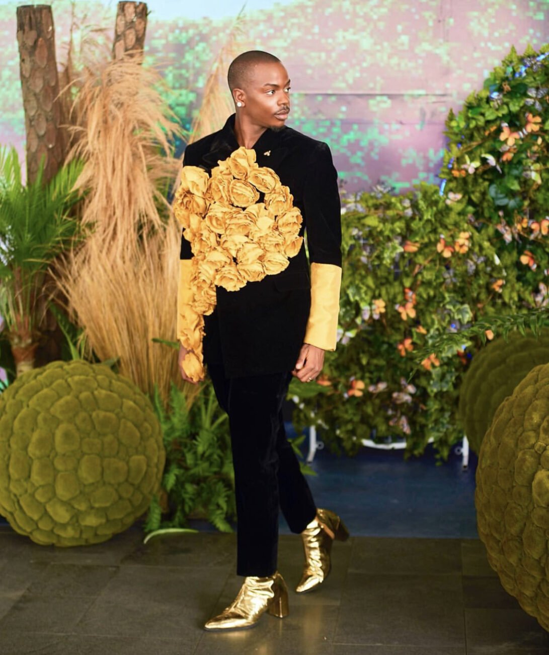 Enioluwa Adeoluwa in a black suit with yellow roses attached to the front, and yellow sleeves, alongside gold colored boots.