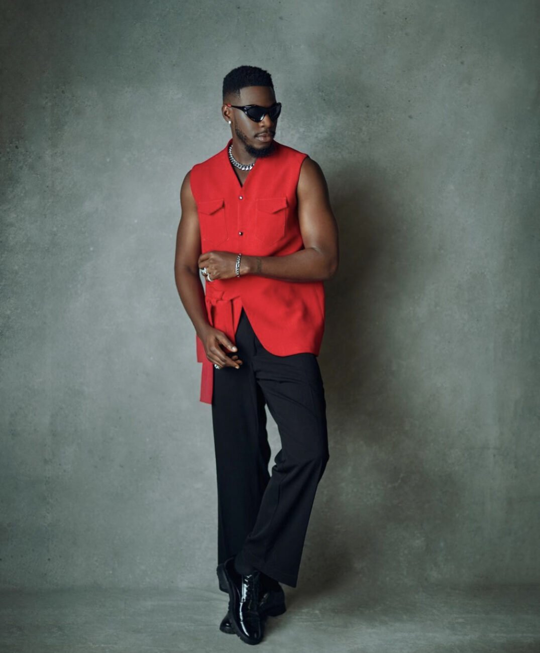 BBNaija’s Soma in a sleeveless red shirt with multiple pockets, black trousers and shiny black shoes. 