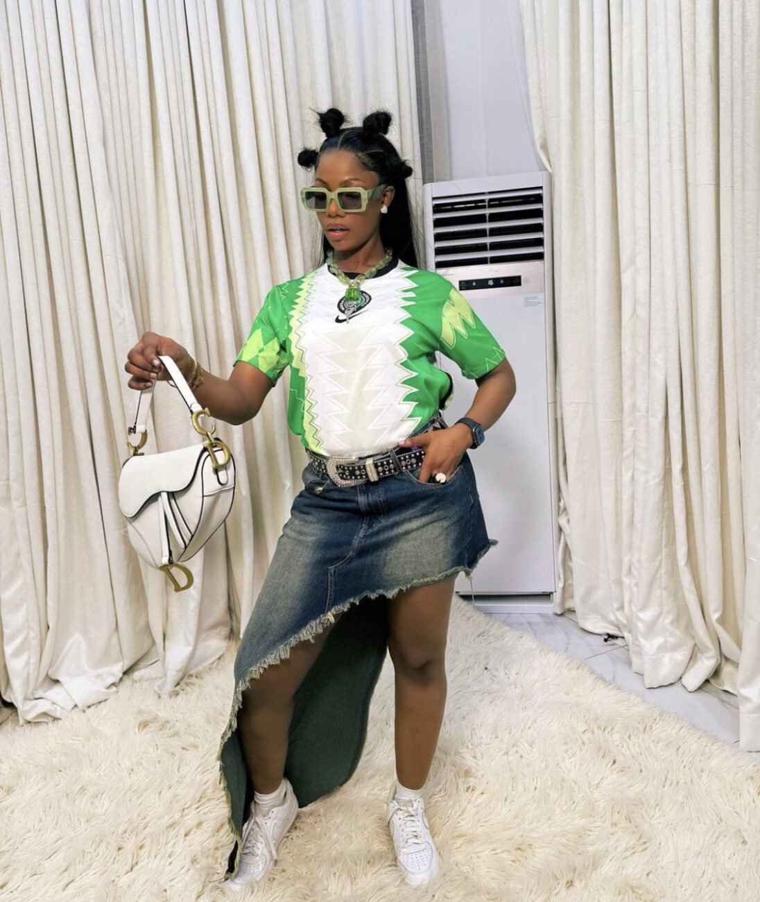 Tacha in a Nigerian jersey, paired with a denim skirt with an asymmetrical hemline, and white sneakers, alongside a white purse.